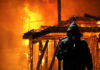 Hooded figure holding match in front of burning building