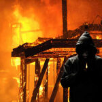 Hooded figure holding match in front of burning building