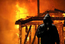 Hooded figure holding match in front of burning building