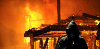 Hooded figure holding match in front of burning building