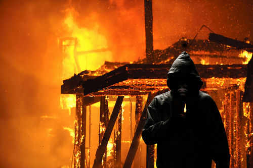 arsonist244348039 Hooded figure holding match in front of burning building