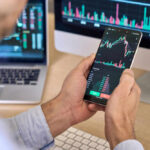 China CUTS Off Foreign Investments – What’s Next? Person checking stock market graphs on phone and monitors.