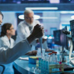 Scientist using pipette at lab bench with test tubes and monitors