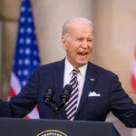 elderly man in suit speaking at podium with microphones