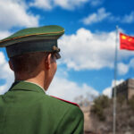 Soldier in a green military uniform facing the Great Wall of China with the Chinese flag in the background