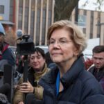 woman in glasses speaking to reporters outdoors