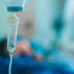 Missed Warning Signs Ended In Tragedy Close-up of an IV drip in a hospital setting with a patient in the background