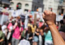A Star-Studded Protest Hits A Roadblock Raised fist in foreground at a crowded street protest