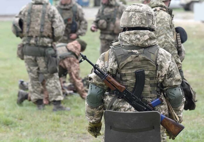Soldier in camouflage with rifle and body armor stands near a group in a field